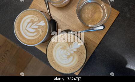 Zwei Tassen frisch zubereiteter Latte mit wunderschöner Latte Art, serviert auf einem Holztablett in einem Café. Die Schaummuster sorgen für einen perfekten Kaffeemoment. Stockfoto