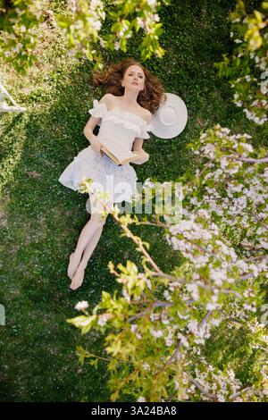 Romantische Frau in einem weißen Kleid, die auf grünem Gras unter Kirschblüten liegt, in Frühlingssonne ein Buch liest, traumhafte und friedliche Szene. Stockfoto