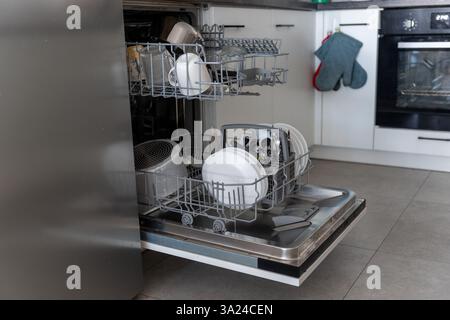 Die moderne Küche mit weißen Schränken verfügt über einen offenen Geschirrspüler mit sauberem Geschirr, Besteck und Kochutensilien. Konzept der Haushaltsgeräte, Küchenorganisation Stockfoto
