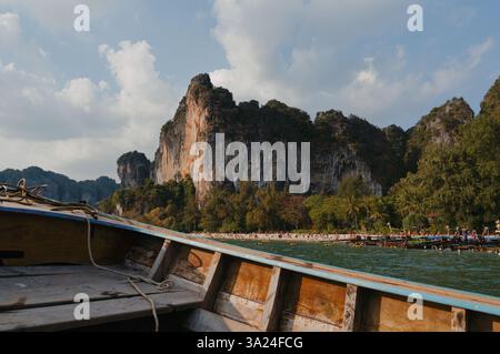 Ein traditionelles Holzboot gleitet in Richtung Railay Beach, wo Kalksteinklippen über goldenen Sand und türkisfarbenem Wasser ragen Stockfoto
