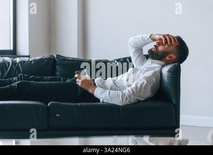 Kopfschmerzen haben, auf dem Sofa liegen. Verärgerter junger Mann Trinker Alkohol ist drinnen. Stockfoto