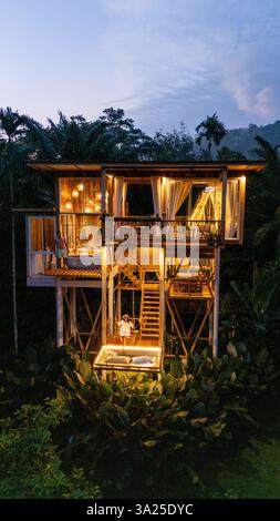 Das wunderschön gestaltete Baumhaus liegt inmitten von lebendigem Grün und leuchtet warm, wenn die Dämmerung einbricht. Gäste genießen ruhige Momente auf der Terrasse, umgeben von Natur Ruhe in Thailand oder Bali. Stockfoto