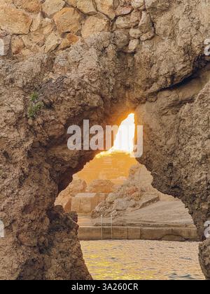 Eine Felsformation mit einem Loch darin und einer Sonne, die durchscheint. Die Sonne untergeht und das Licht schafft eine warme, friedliche Atmosphäre Stockfoto
