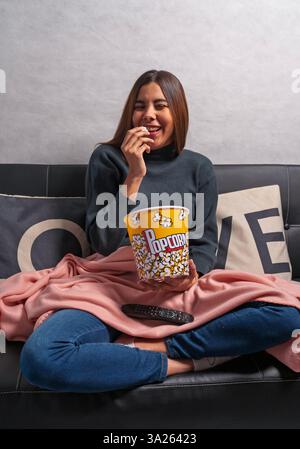 Glückliche junge Frau lacht, während sie Popcorn isst und einen Film auf dem Sofa zu Hause ansieht Stockfoto