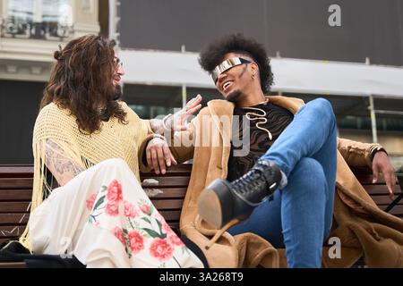 Ein paar androgyne nicht-binäre Leute, die moderne Kleidung tragen, entspannen sich auf einer Bank, plaudern und lächeln Stockfoto