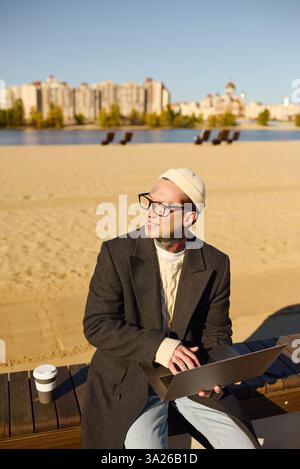 Gutaussehender Mann mit Tattoos lächelt, während er am Strand sitzt und das warme Wetter und die Landschaft genießt. Stockfoto