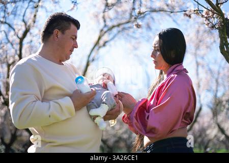 Lateinische Eltern kümmern sich um ihr neugeborenes Baby in einem blühenden Obstgarten und genießen einen sonnigen Frühlingstag Stockfoto