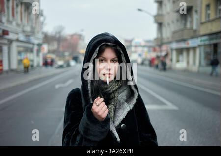 Eine Frau in einem Kapuzenmantel steht auf einer verlassenen Straße und hält eine brennende Zigarette. Berlin, Deutschland Stockfoto