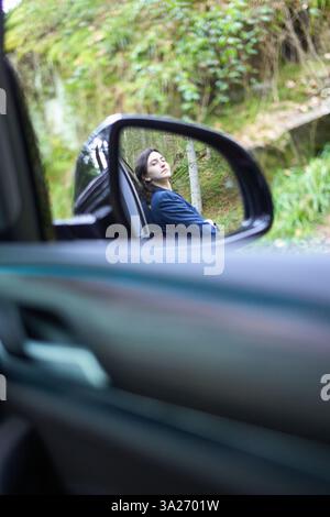 Frau, die sich an ihr Auto lehnt, von innen gesehen durch den Seitenspiegel, mit grünem Waldhintergrund. Stockfoto