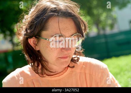 Junge Frau mit Brille, die draußen im Sonnenlicht sitzt, ein pfirsichfarbenes Hemd trägt, mit Augen nach unten. Stockfoto