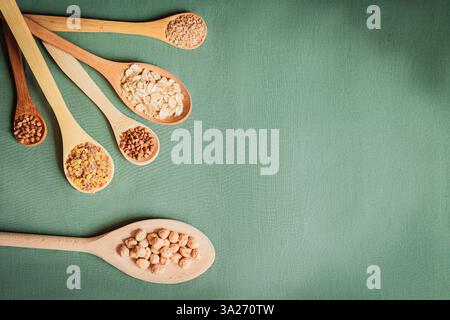 Auswahl von Körnern und Getreide in Holzlöffeln, die auf einer strukturierten grünen Oberfläche nach außen strahlen. Frisches, biologisches Lebensmittelkonzept mit Platz für Text Stockfoto