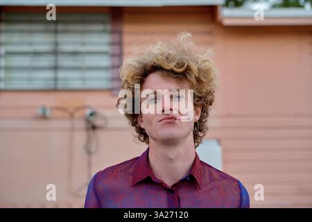 Junger Mann mit lockigen Haaren, der ein rotes Hemd trägt und vor einem pfirsichfarbenen Gebäude raucht. Miami, FL, USA Stockfoto
