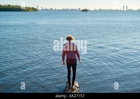 Eine Person mit lockigen Haaren steht auf einem Felsen im Wasser mit Blick auf die entfernte Skyline der Stadt Miami, FL, USA Stockfoto