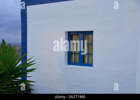 Blau gerahmtes Fenster auf einer weißen Stuckwand mit einer grünen Pflanze im Vordergrund. Lanzarote, Spanien Stockfoto