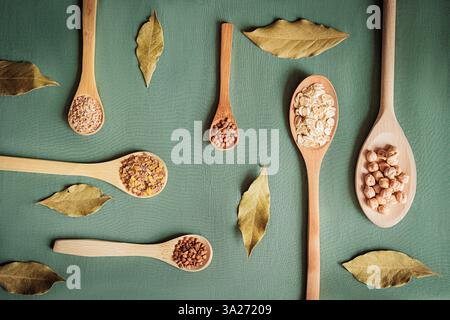 Flache Lage von Körnern, Hülsenfrüchten und Getreide in Holzlöffeln mit Lorbeerblättern auf grünem Hintergrund. Bio-Lebensmittel und gesunde Ernährung Konzept, perfekt für Stockfoto