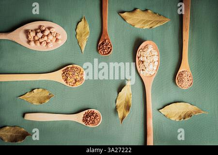 Flache Lage von Körnern, Hülsenfrüchten und Getreide in Holzlöffeln mit Lorbeerblättern auf grünem Hintergrund. Bio-Lebensmittel und gesunde Ernährung Konzept, perfekt für Stockfoto