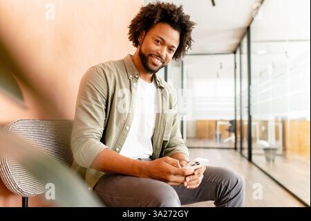 Portrait von lächelnden afroamerikanischen Mann auf dem Sofa im Büro sitzen, mit Handy für die Überprüfung der sozialen Netze, Eingabe von Nachricht für Familie, Freundin, Geschäftspartner, Ansichten Fotos, Videos Stockfoto