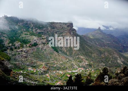 Neblige Berglandschaft mit zerklüfteten Klippen und einem kleinen Dorf im Tal darunter. Gran Canary, Spanien Stockfoto