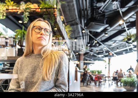 Stilvolle junge Frau mit Brille und langen blonden Haaren, die in einem modernen Café mit urbanem Dekor sitzt und zur Seite blickt. Entspannte Atmosphäre mit natürlichem Ambiente Stockfoto