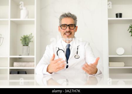 Ein Oberarzt mit grauen Haaren und einem warmen Lächeln macht eine Gesprächsgeste, als ob er eine Diagnose oder einen medizinischen Rat vor dem Hintergrund eines modernen Klinikinnenraums erklärt. Telemedizin Stockfoto