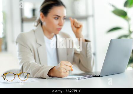 Unfokussierte, frustrierte Geschäftsfrau in einem beigefarbenen Blazer sitzt am Schreibtisch und ballt ihre Faust, während sie einen Laptop-Bildschirm mit einem fokussierten Ausdruck ansieht, der unzufrieden und wütend aussieht Stockfoto