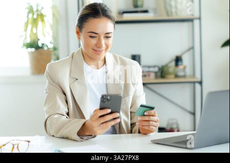 Die lächelnde kaukasische Geschäftsfrau im beigefarbenen Blazer hält ein Smartphone und eine Kreditkarte, während sie am Schreibtisch eine Online-Zahlung tätigt, Finanzen verwaltet oder über eine mobile Banking-App einkaufen kann Stockfoto