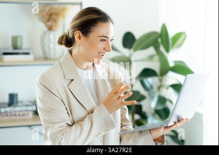 Die aufgeregte emotionale kaukasische Geschäftsfrau in einem eleganten Kleiderständer im Büro, hält einen Laptop in einer Hand und gibt mit einer anderen ausdrucksstarke Gesten, bekam unerwartete gute Nachrichten Stockfoto