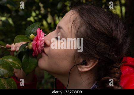 Eine Frau in einer roten Jacke hält und riecht sanft eine rosafarbene Kamelienblume mit geschlossenen Augen, die die friedliche und sensorische Schönheit der Natur umschmeichelt. Stockfoto