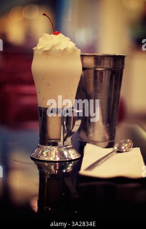 Ein köstlicher Milchshake mit Schlagsahne und einer Kirsche, serviert in einem klassischen Glas mit einem Metallshaker an der Seite. Die Einrichtung ist ein Diner mit einem Stockfoto