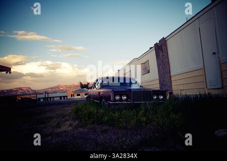 Ein altes, rostiges Auto parkt auf bewachsenem Gras in der Nähe eines Wohnwagens, mit Bergen im Hintergrund unter einem bunten Himmel. Stockfoto