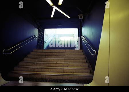 Blick auf eine Treppe, die zu einem hellen Ausgang führt, eingerahmt von dunklen Wänden und moderner Beleuchtung. Die Treppen sind aus Beton und die Atmosphäre ist städtisch und Stockfoto