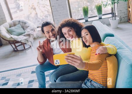 Eine Gruppe verschiedener Freunde macht ein Selfie auf der Couch in einem hellen Wohnzimmer, in dem Wärme, Freude und eine fröhliche Wochenendatmosphäre zum Ausdruck kommen. Stockfoto