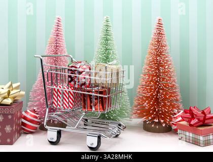 Nahaufnahme eines kleinen Einkaufswagens mit bunten Geschenken vor bunten Weihnachtsbäumen und Geschenken, grüne, abgestreifte Wand dahinter. Stockfoto