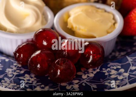 Ein Teller mit roten Kirschen und weißer Clotted Cream liegt auf einem blau-weißen Teller. Die Kirschen sind so angeordnet, dass sie aussehen, als wären sie bereit Stockfoto