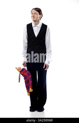 Ein Mann in schwarzer Weste und weißem Hemd steht mit einer Puppe, die einem Affen ähnelt, in einem karierten Outfit. Der Hintergrund ist schlicht weiß, wodurch das su betont wird Stockfoto