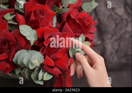 Die Hände der Floristin berühren Rosenblätter und arrangieren einen Strauß roter Rosen mit Eukalyptuszweigen auf schwarzem, dunklem Relief-Hintergrund Stockfoto