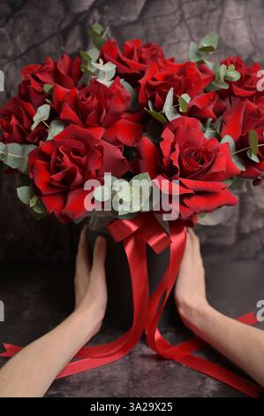 Frauenhände halten einen Strauß roter Rosen mit Eukalyptus in Black Box und rotem Band. Schwarzer dunkler Reliefhintergrund. Blumenarrangements Stockfoto