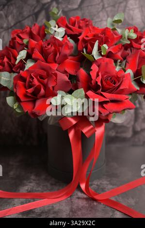 Strauß aus roten Rosen mit Eukalyptus in Black Box und rotem Band. Schwarzer dunkler Reliefhintergrund. Blumenarrangements. Tapete, Hintergrund, Cover Stockfoto