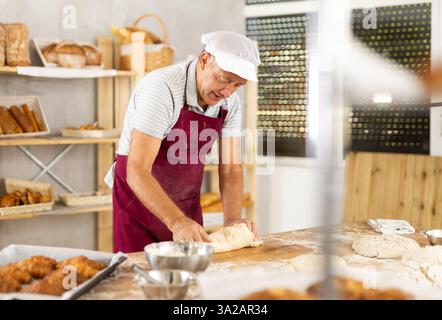 Senior Mann arbeitet in der Bäckerei als Bäcker, Knete Teig, arbeitet mit Mehl. Stockfoto