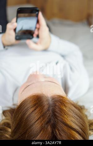 Ein rothaariges Mädchen liegt auf dem Bett neben einem offenen Laptop, während es am Telefon telefoniert, was eine entspannte und ungezwungene Szene schafft. Stockfoto