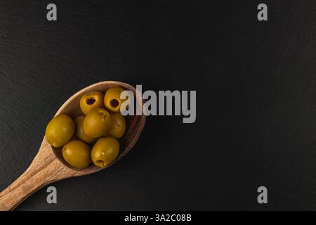 Ein Holzlöffel fasst mehrere grüne Oliven, die ordentlich angeordnet sind. Der dunkle Hintergrund verstärkt die natürlichen Farben der Oliven und schafft ein ansprechendes Erscheinungsbild. Stockfoto