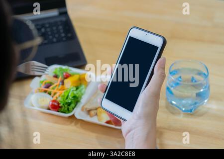 Nahaufnahme der Frau essen Mittagessen mit gesunder Nahrung, wenn sie Smartphone. Rückansicht, selektiver Fokus. Stockfoto