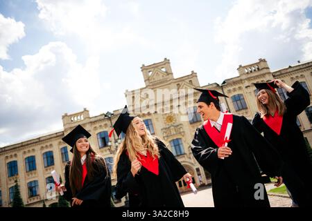 Vier junge Absolventen feiern vor einem prunkvollen Universitätsgebäude, halten Diplome und tragen traditionelle akademische Kleidung. Stockfoto