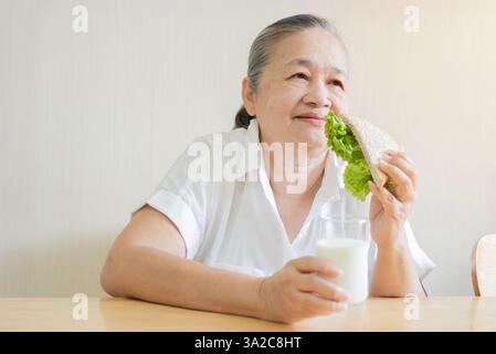 Glückliches Porträt einer älteren asiatischen Frau, die auf einem Holztisch sitzt und Gemüse-Sandwich und Milch isst. Kopierbereich, selektiver Fokus. Stockfoto