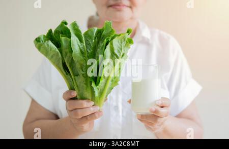 Frischer Cos-Salat und Milchglas in den Händen einer intelligenten gesunden älteren Frau beim Stehen über hellbeigem Hintergrund. Selektiver Fokus. Stockfoto