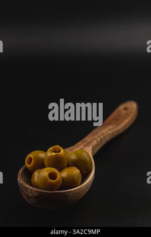 Ein Holzlöffel fasst mehrere grüne Oliven und zeigt ihre glatte Textur und einzigartige Form. Der dunkle Hintergrund hebt die natürliche Farbe der Oliven hervor. Stockfoto