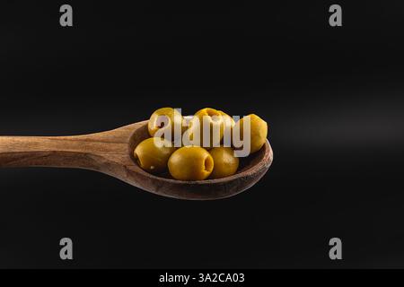 Ein Holzlöffel hält mehrere grüne Oliven und hebt ihre glatten Oberflächen hervor. Der dunkle Hintergrund unterstreicht die lebendige Farbe und Textur der Oliven. Stockfoto