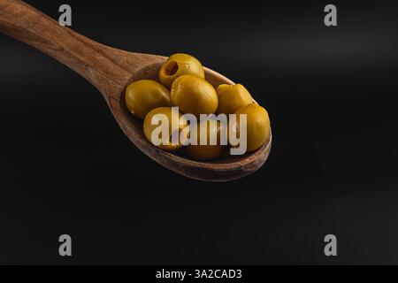 Ein Holzlöffel fasst mehrere kugelförmige grüne Oliven, die ihre glatte Textur und leuchtende Farbe vor einem dunklen Hintergrund zeigen. Die Komposition Highli Stockfoto