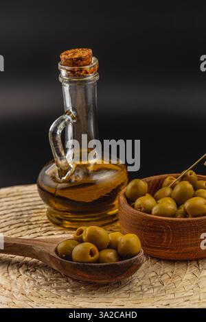 Frisch geerntete grüne Oliven werden in eine Holzschale neben einer mit Olivenöl gefüllten Glasflasche gelegt, die auf einer gewebten Matte vor dunklem Hintergrund steht. Stockfoto