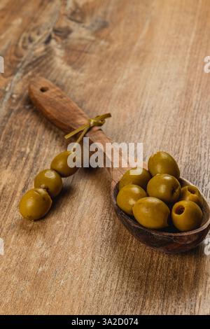 Ein Holzlöffel hält mehrere grüne Oliven und zeigt ihre glänzende Textur. Ein paar Oliven sind in der Nähe auf einem rustikalen Holztisch verstreut. Die Einstellung exu Stockfoto